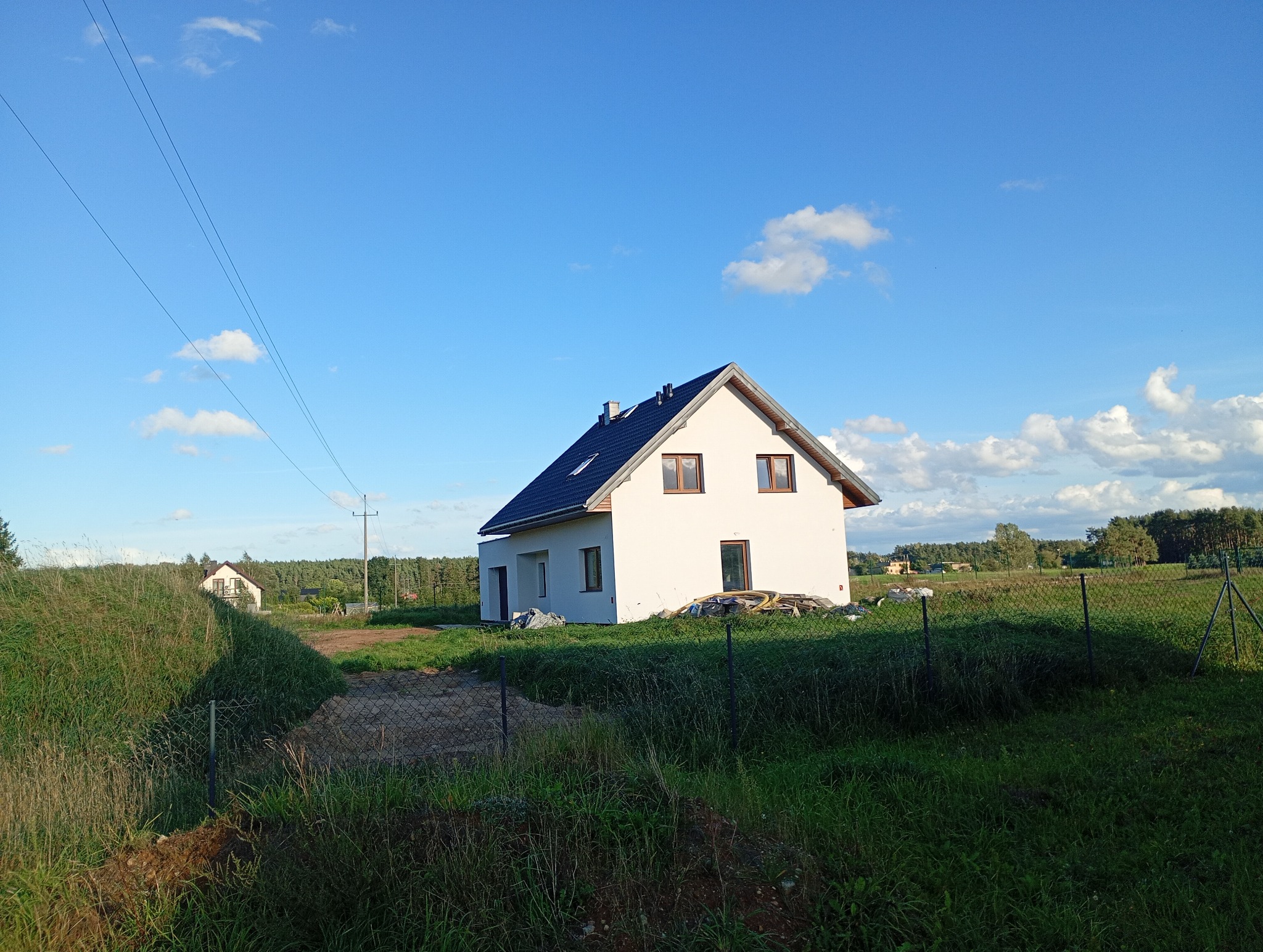 Nowy, biały dom jednorodzinny z ciemnym dachem na tle błękitnego nieba i zieleni. Widoczne elementy wykończeniowe i teren wokół domu.
