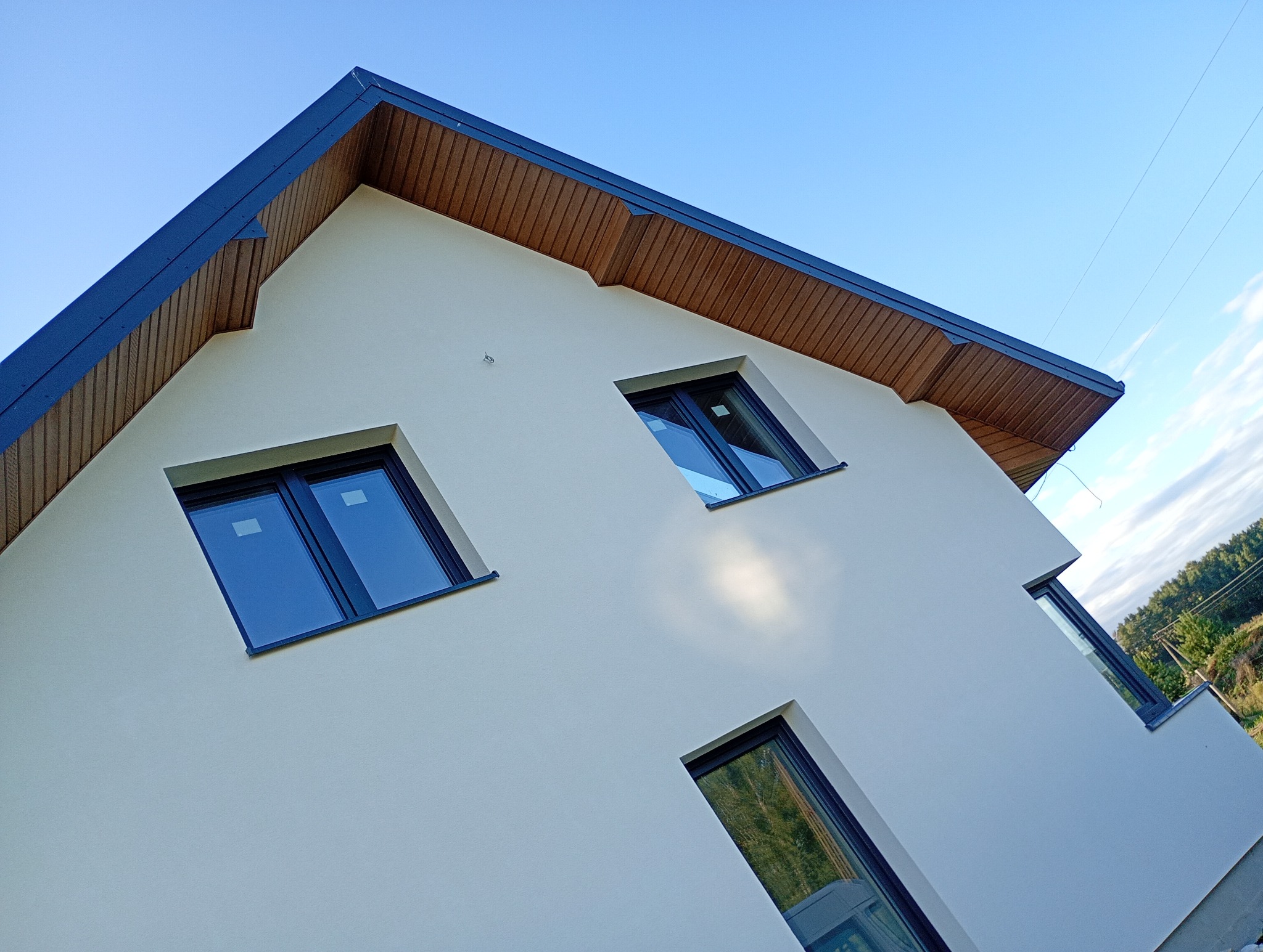 Elewacja domu jednorodzinnego z drewnianą podbitką, widziana z dołu pod kątem. Biała ściana z trzema oknami o czarnych ramach odbija błękit nieba.