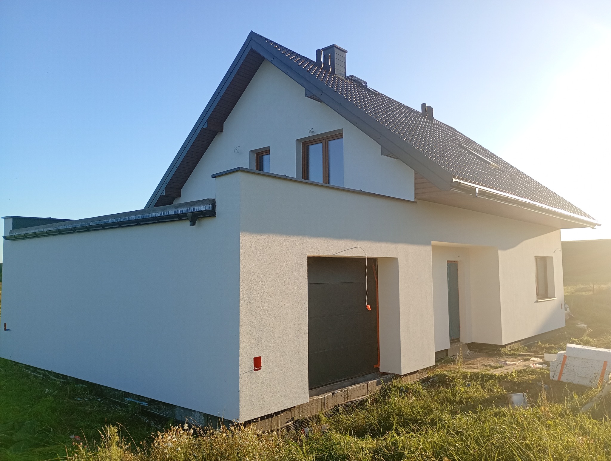 Nowoczesny dom jednorodzinny z garażem, wykończony jasnym tynkiem i ciemną dachówką, w otoczeniu zieleni. Widoczne okna z brązowymi ramami i elementy instalacji.