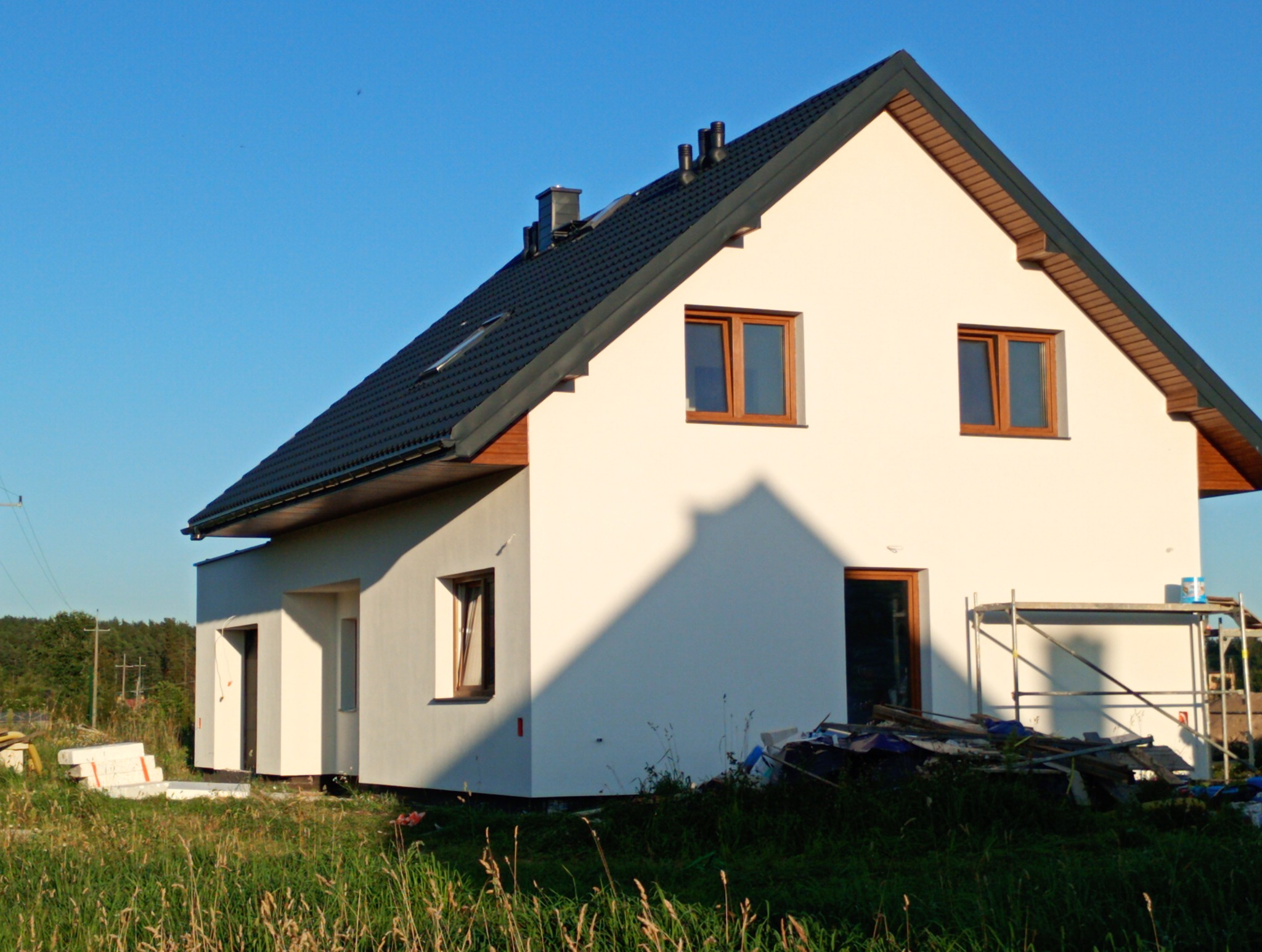 Nowoczesny dom jednorodzinny z czarną dachówką i białymi ścianami, w otoczeniu zieleni. Widoczne rusztowanie i materiały budowlane.