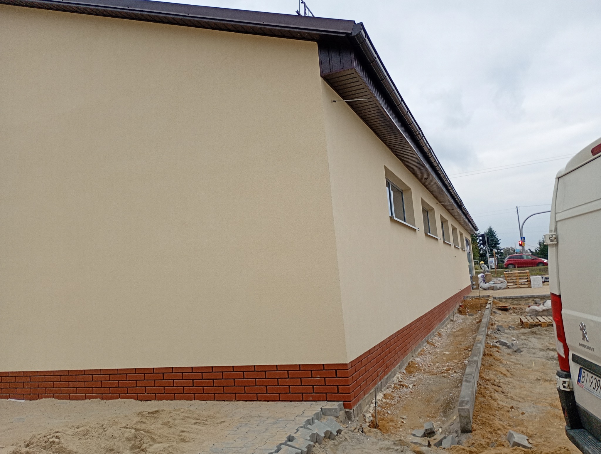 Elewacja budynku po remoncie z ceglaną podmurówką i beżową ścianą. Widoczne okna, fragment chodnika i samochód dostawczy. Prace wykończeniowe w toku.