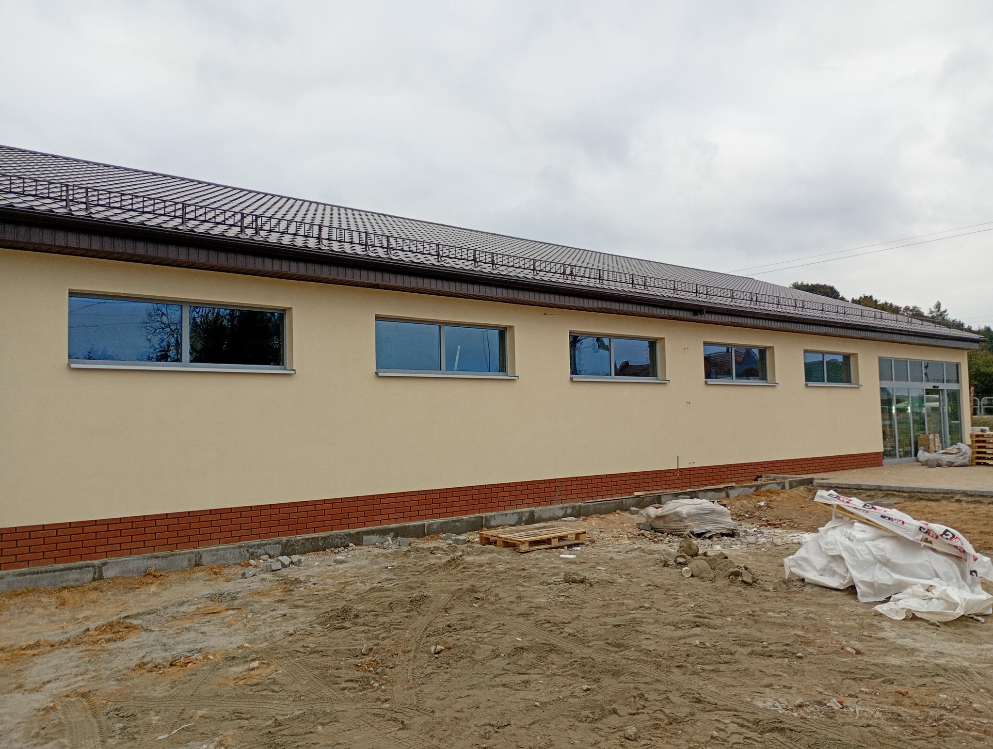 Elewacja budynku po remoncie z beżową ścianą, brązową cegłą i ciemnymi oknami. Na pierwszym planie teren budowy z materiałami.