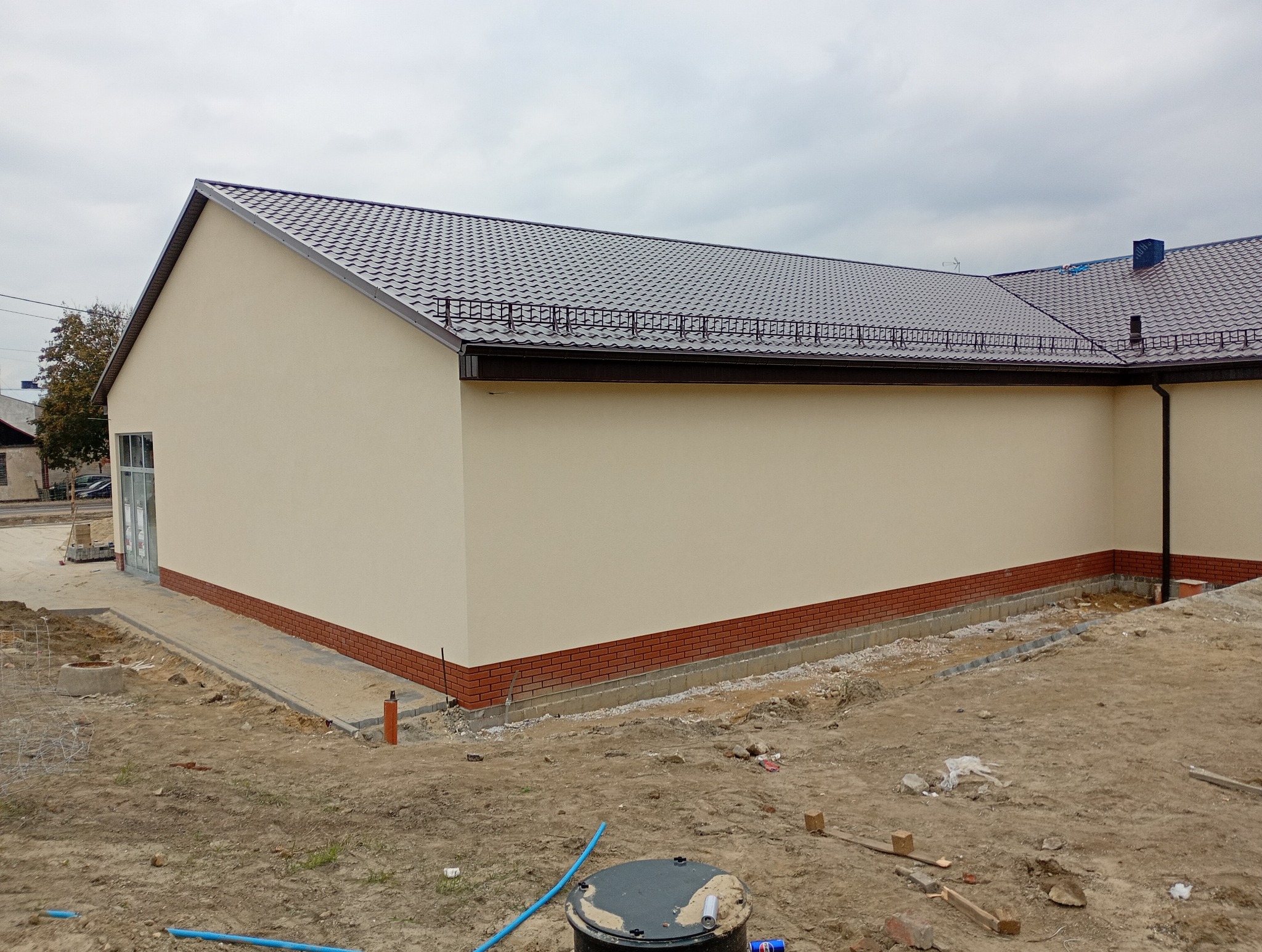 Elewacja budynku z beżową ścianą i czerwoną cegłą u podstawy, ciemny dach z metalowej blachy. Teren wokół budynku w trakcie prac wykończeniowych.