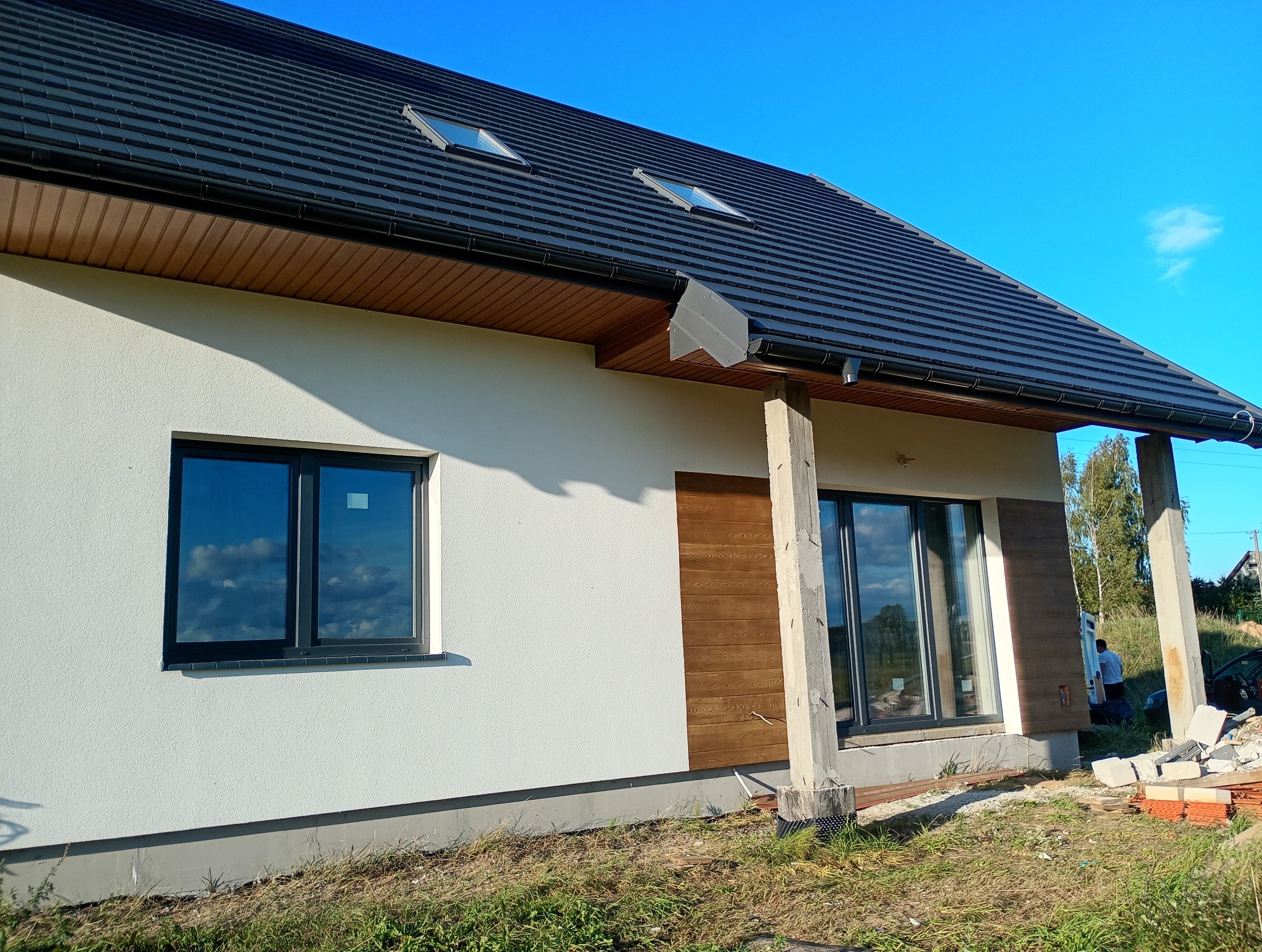 Elewacja domu jednorodzinnego z czarną dachówką i drewnianą okładziną. Widoczne okna z odbiciem nieba i fragment terenu z gruzem.