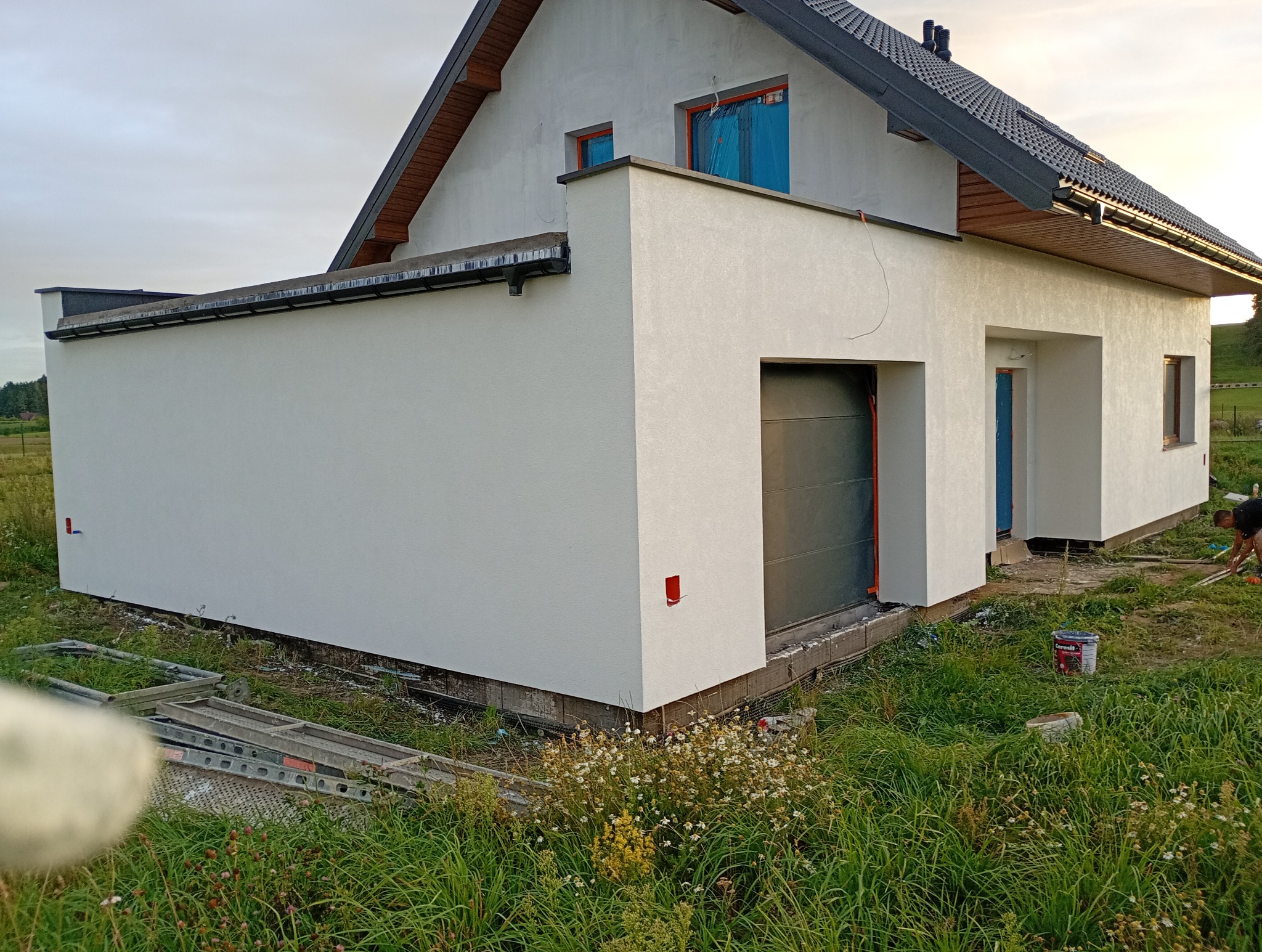 Otynkowany dom jednorodzinny z garażem, świeżo po malowaniu elewacji na biało. Widoczne okna zabezpieczone folią, drabina i wiadro z farbą. Prace wykończeniowe.