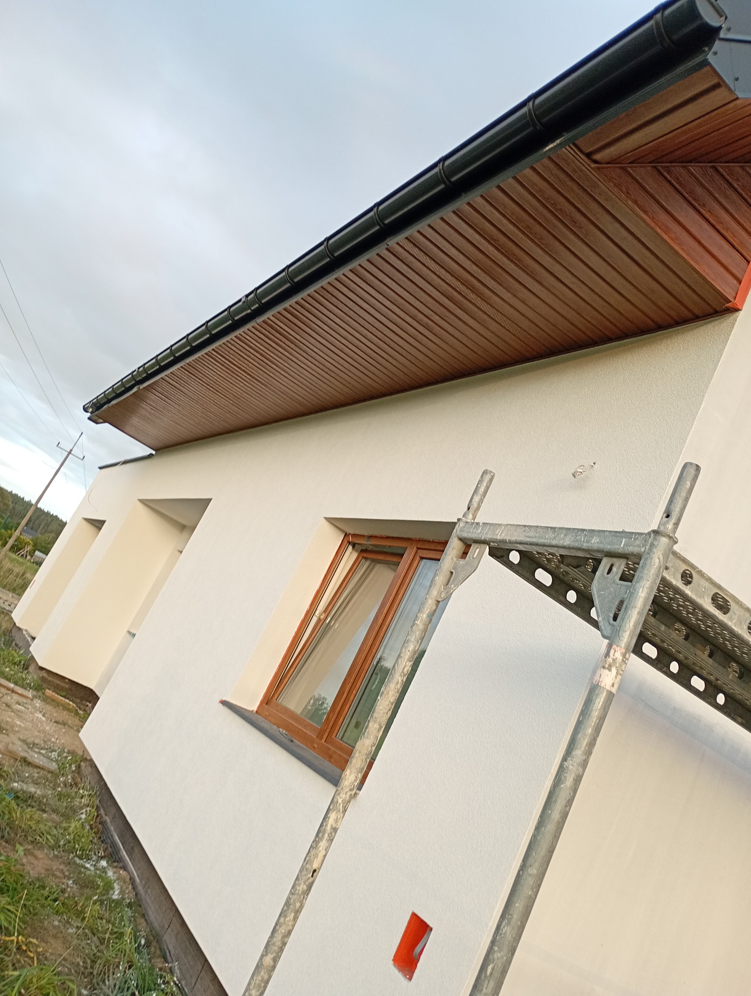 Elewacja domu jednorodzinnego po malowaniu, widoczne rusztowanie, okno z brązową ramą, podbitka dachowa w kolorze drewna i czarna rynna.