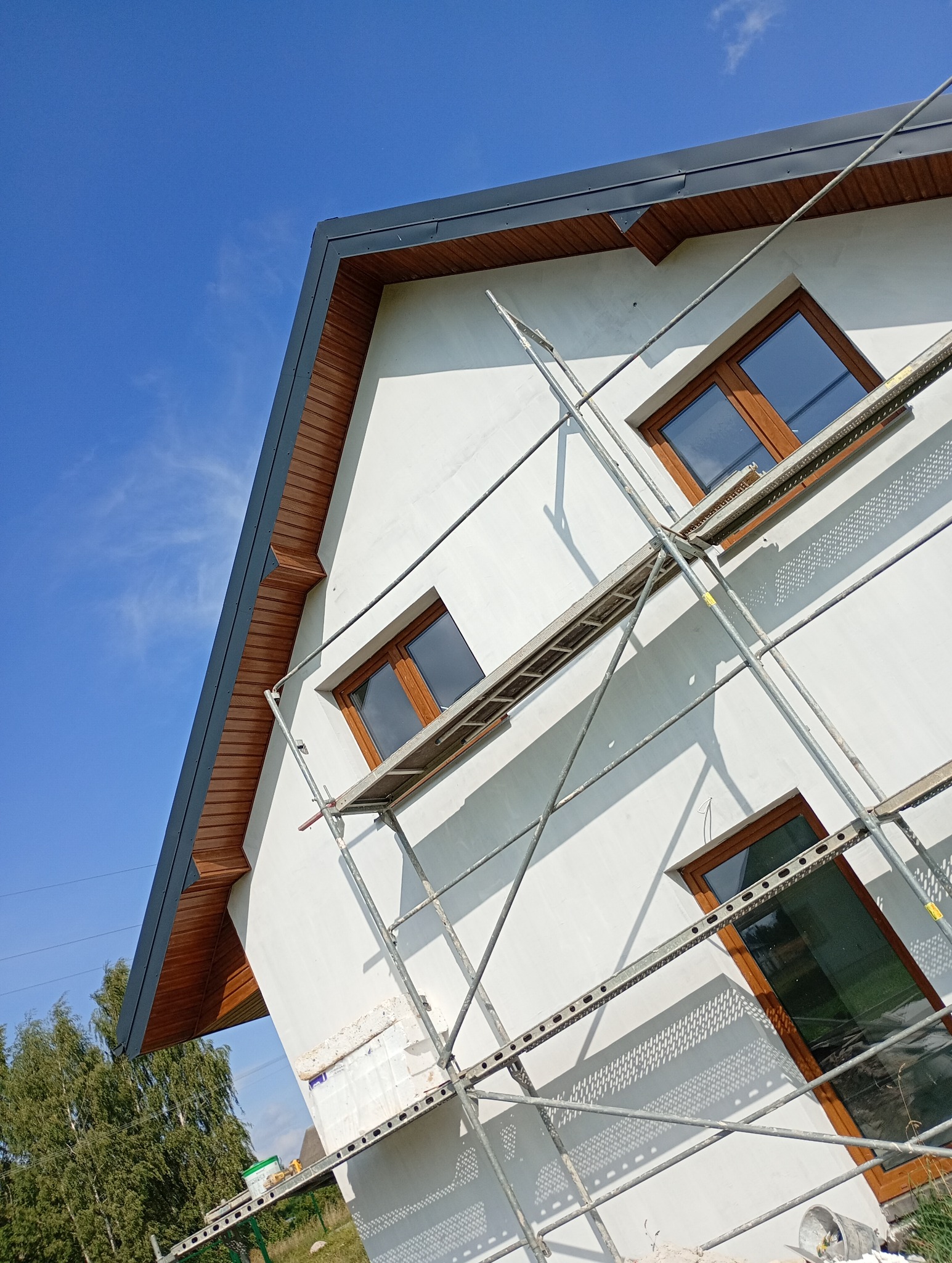 Dom jednorodzinny w trakcie prac wykończeniowych elewacji, widoczne rusztowanie i fragment ocieplenia. Ujęcie z dołu, błękitne niebo w tle.