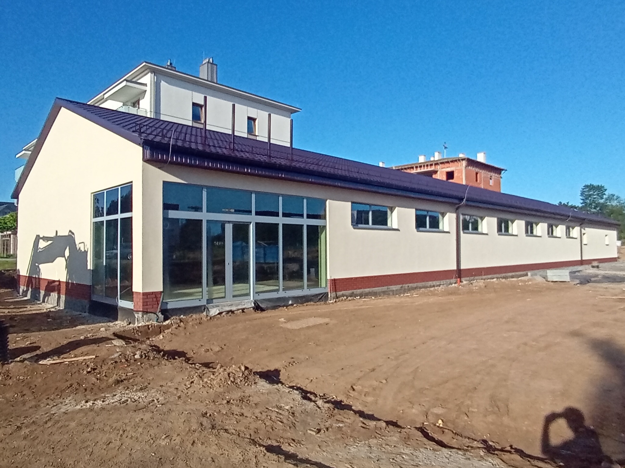 Budynek parterowy z ciemnym dachem i dużymi oknami, z widocznym fragmentem wykopu ziemnego przed budynkiem. W tle budynek mieszkalny i niebo.