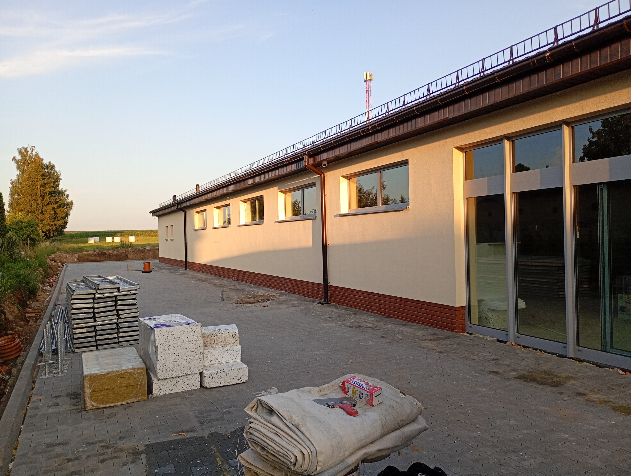 Budynek w trakcie prac wykończeniowych: ocieplenie, materiały budowlane, narzędzia na zewnątrz. Widoczne okna i elewacja w ciepłym świetle.