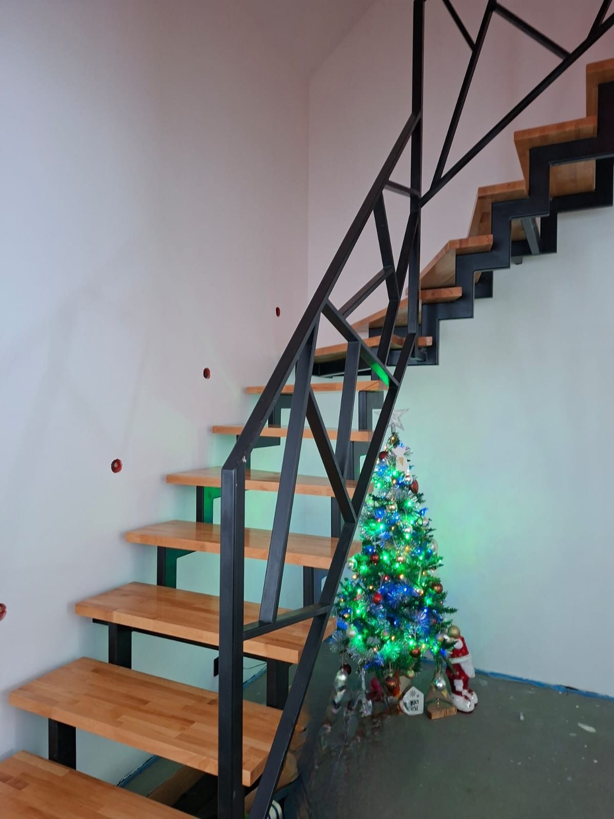 Nowoczesne schody z drewnianymi stopniami i czarną, geometryczną konstrukcją metalową, ozdobione świąteczną choinką w tle.