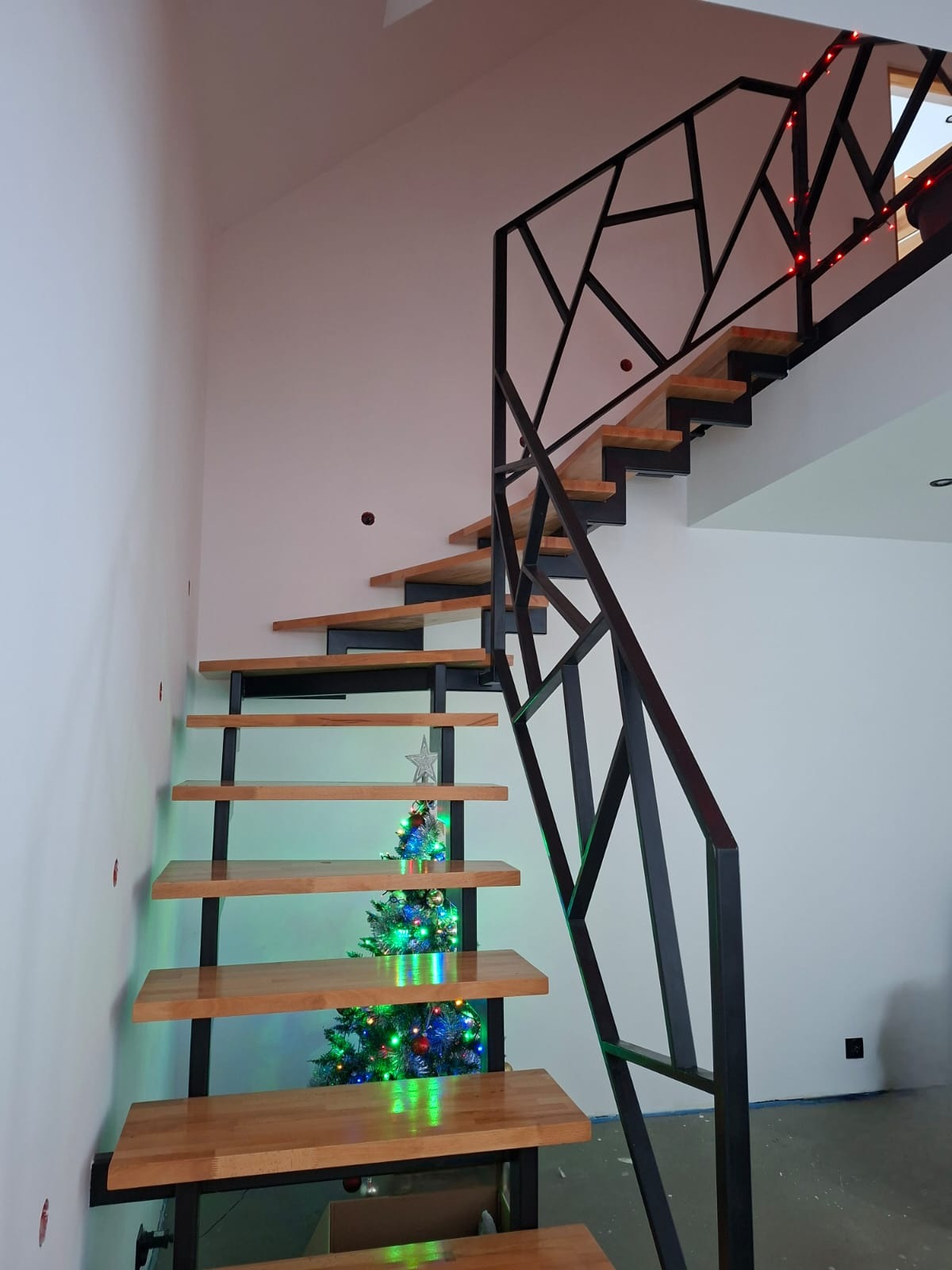 Nowoczesne schody z drewnianymi stopniami i geometryczną, czarną konstrukcją. W tle choinka z lampkami, dodająca świątecznego klimatu wnętrzu.
