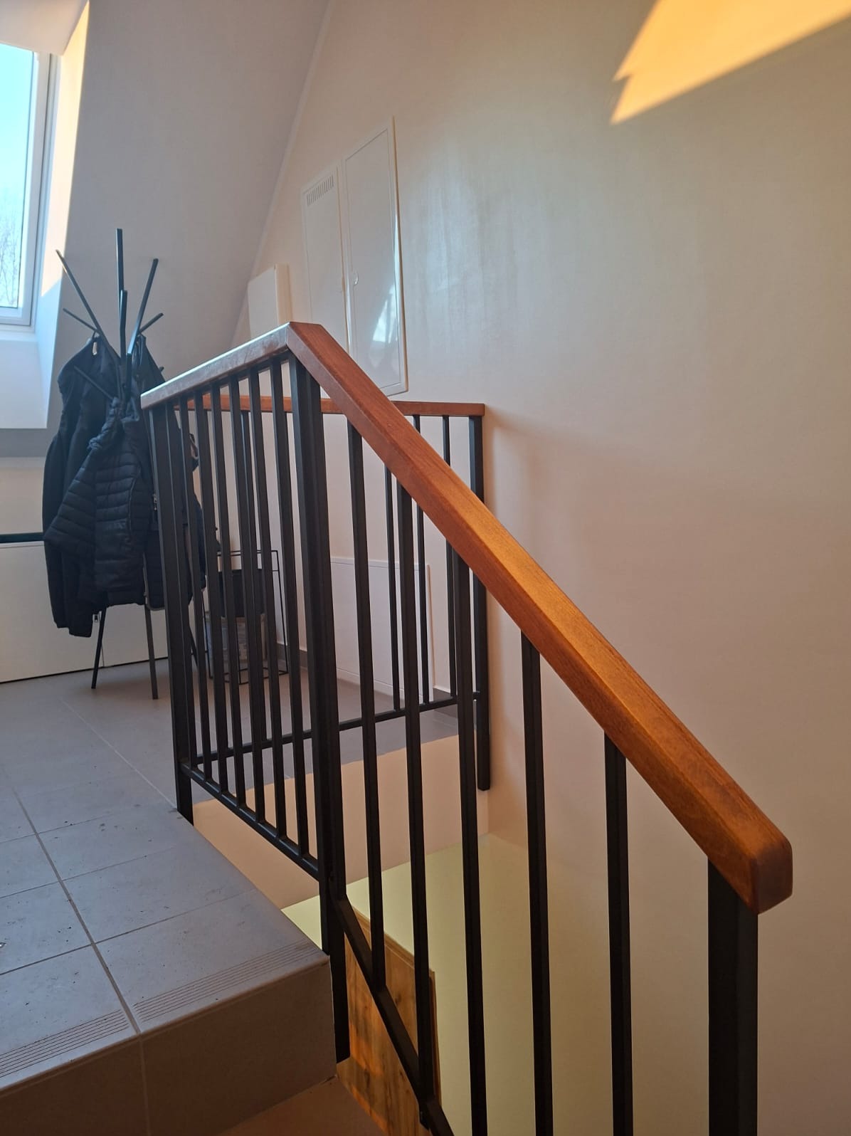 Nowoczesna balustrada schodowa z czarnymi, pionowymi prętami i drewnianą poręczą, oświetlona ciepłym światłem słonecznym w jasnym wnętrzu.