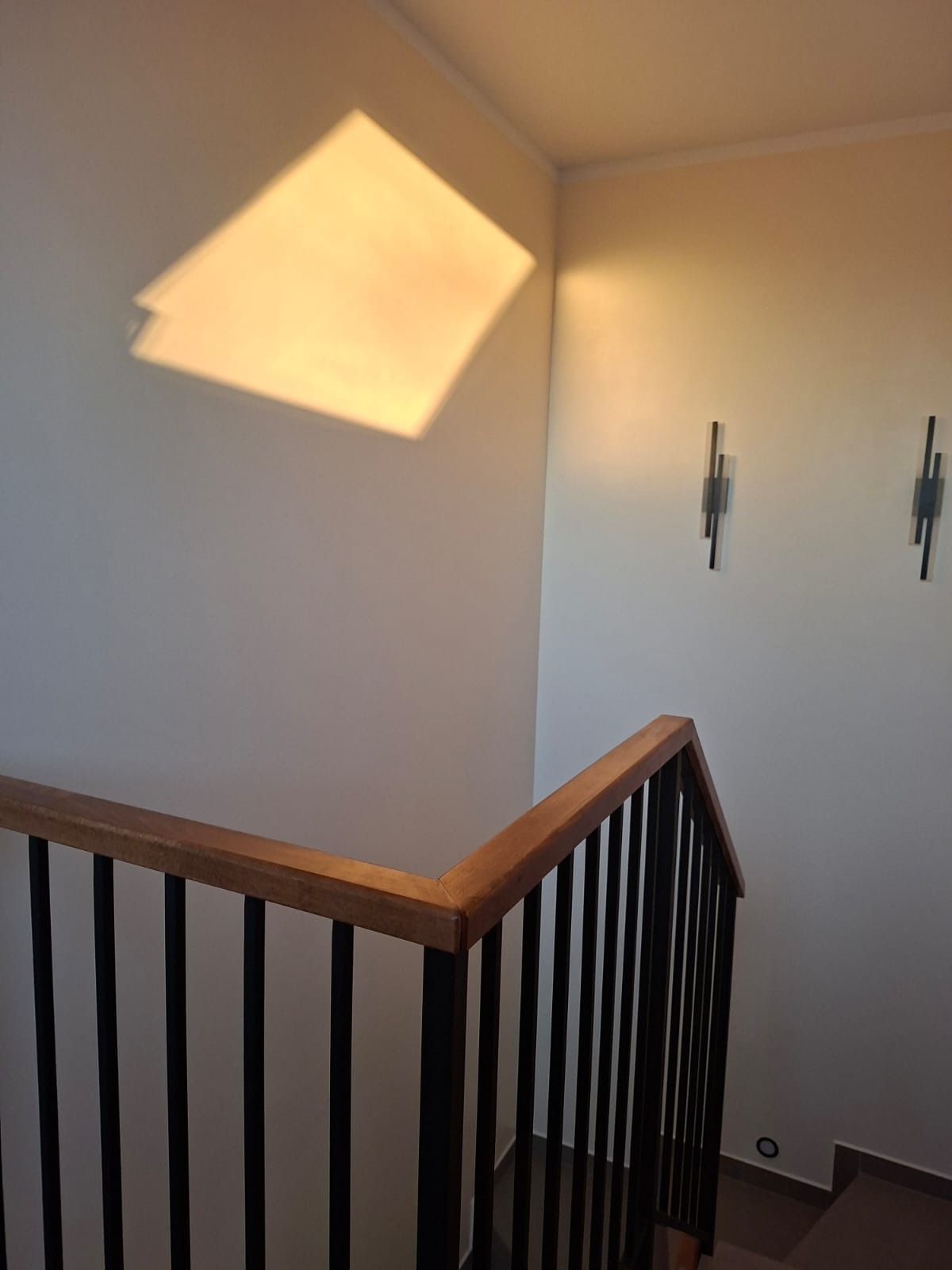 Nowoczesna balustrada schodowa z drewnianym pochwytem i czarnymi, pionowymi elementami, oświetlona ciepłym światłem, z widocznymi minimalistycznymi kinkietami.