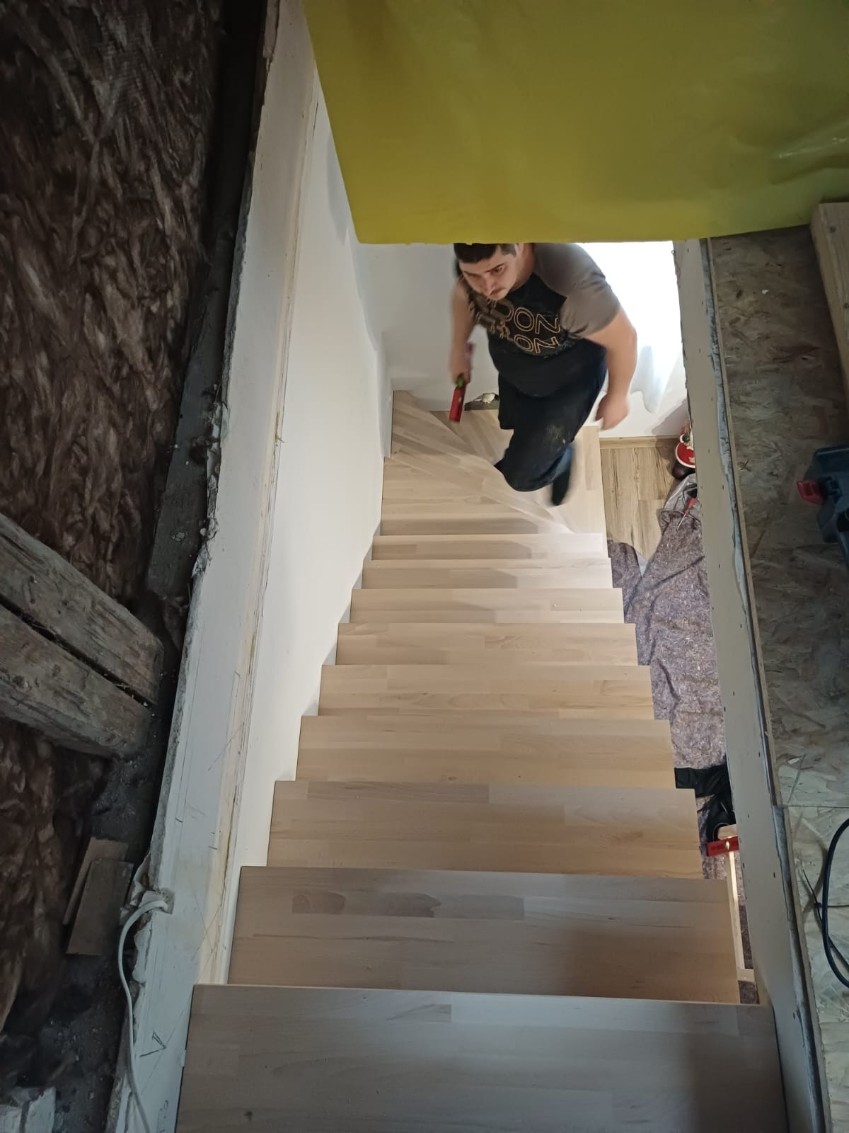 Montaż drewnianych schodów wewnątrz budynku. Widok z góry na schody z jasnego drewna, monter z narzędziem w dłoni. Częściowo wykończone wnętrze.