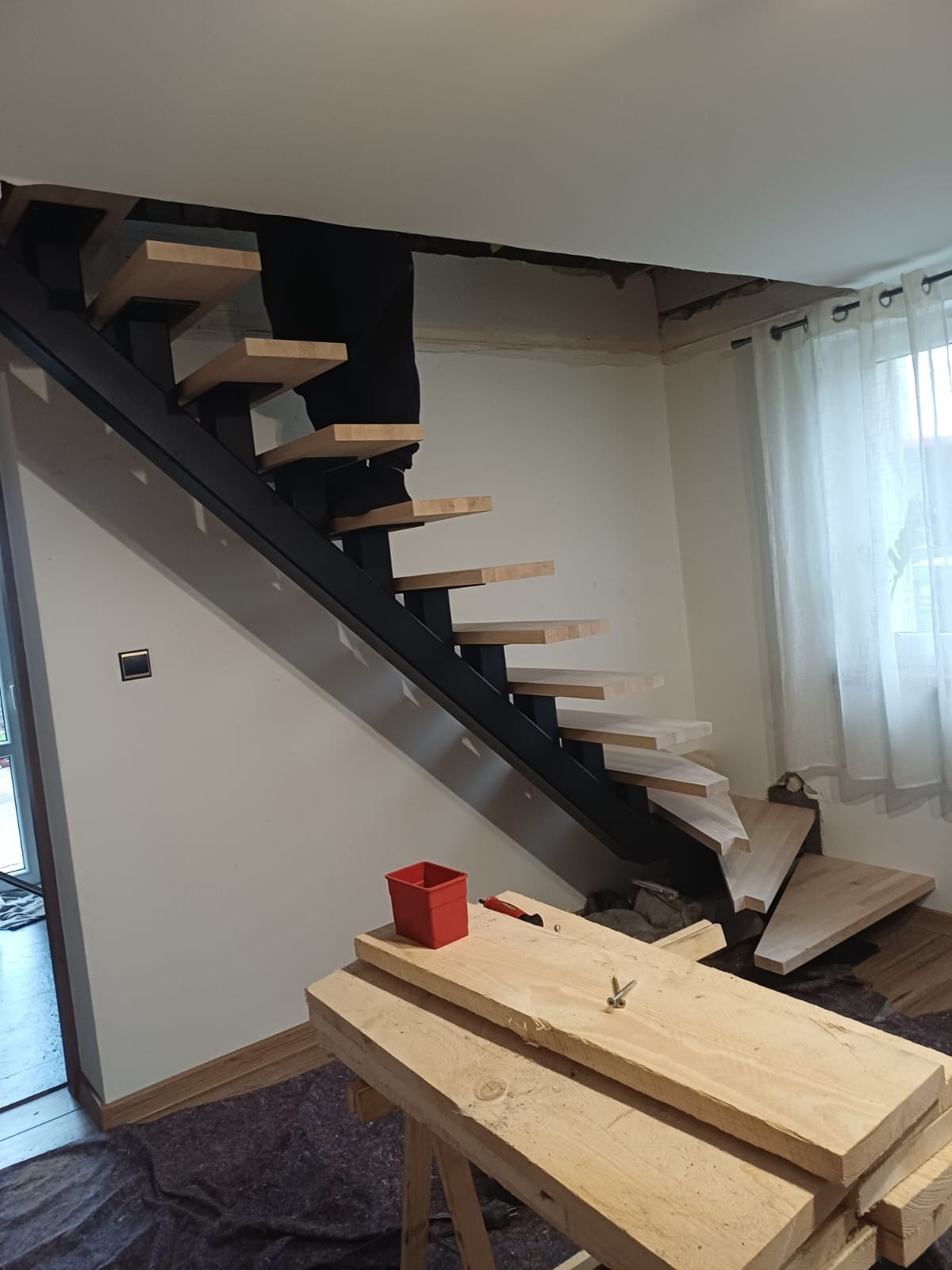 Montaż schodów: czarna konstrukcja metalowa z drewnianymi stopniami, widoczne narzędzia i materiały, etap prac wykończeniowych w jasnym wnętrzu.