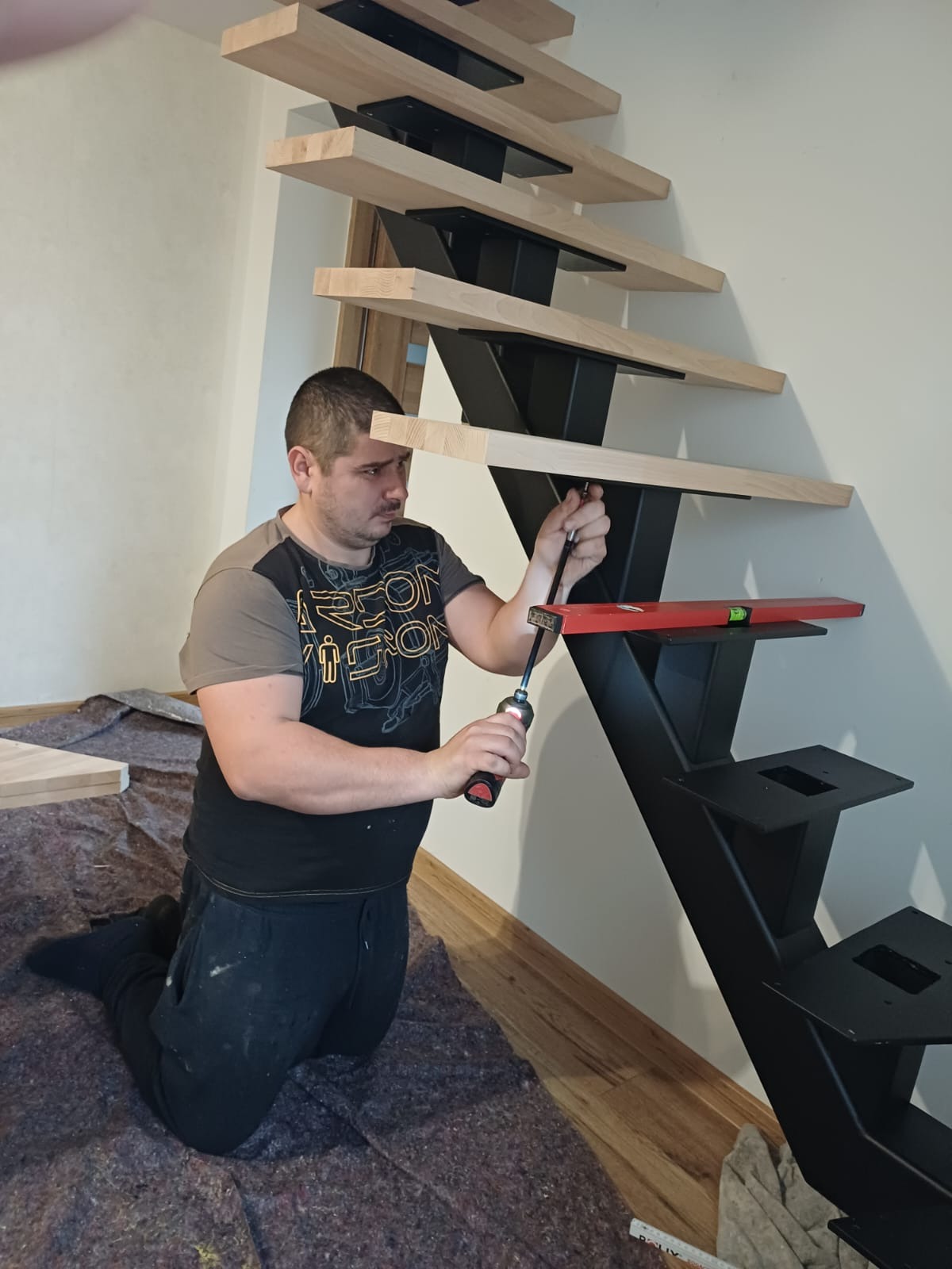 Montaż schodów metalowych z drewnianymi stopniami. Fachowiec z poziomnicą i wkrętakiem przykręca stopień do czarnej konstrukcji. Widoczne zabezpieczenie podłogi.