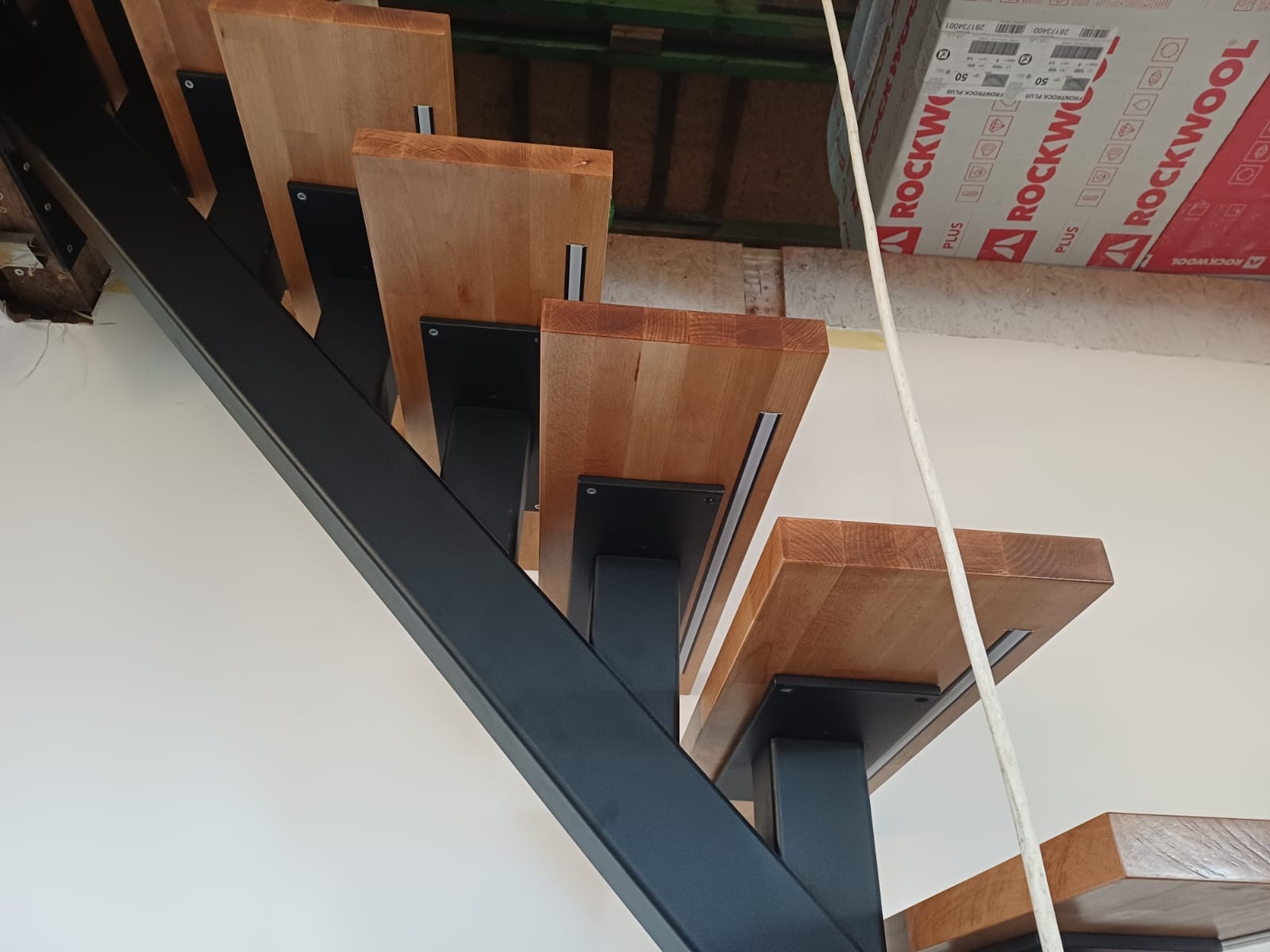 Nowoczesne schody z drewnianymi stopniami i czarną, metalową konstrukcją nośną, widziane z dołu pod kątem, z widocznymi elementami montażowymi.