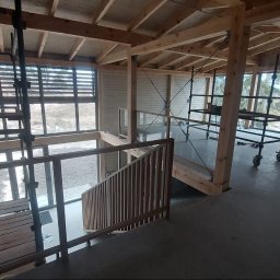 Sneker - Wnętrze domu szkieletowego w trakcie budowy: widoczne drewniane elementy konstrukcyjne, schody z balustradą, rusztowania i okna z żaluzjami. Surowy stan wykończenia.