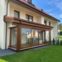 Sneker - Nowoczesna, przeszklona weranda z drewnianą konstrukcją, przylegająca do jasnego domu z brązowym dachem. Widok z zewnątrz na zadbany trawnik i elementy architektury.