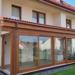 Sneker - Drewniana zabudowa tarasu z przesuwnymi szklanymi drzwiami, dopasowana do elewacji domu z brązową dachówką. Widoczny fragment ogrodu i osoba pracująca na zewnątrz.