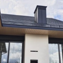 Sneker - Nowoczesny dom z ciemną dachówką i metalowym kominem. Widoczne okna z roletami zewnętrznymi i drewniana podbitka dachowa. Ujęcie z dołu, podkreślające geometrię budynku.