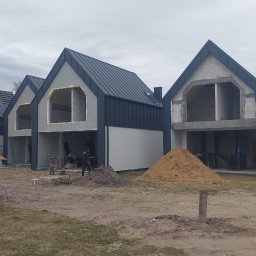 Sneker - Surowe domy w zabudowie szeregowej z ciemnymi dachami z blachy na etapie budowy, widoczne sterty piasku i pracownik w oddali.