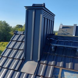 Sneker - Nowoczesny komin wentylacyjny z blachy na dachu krytym ciemną dachówką ceramiczną. Widoczne elementy konstrukcji dachu i niebo.