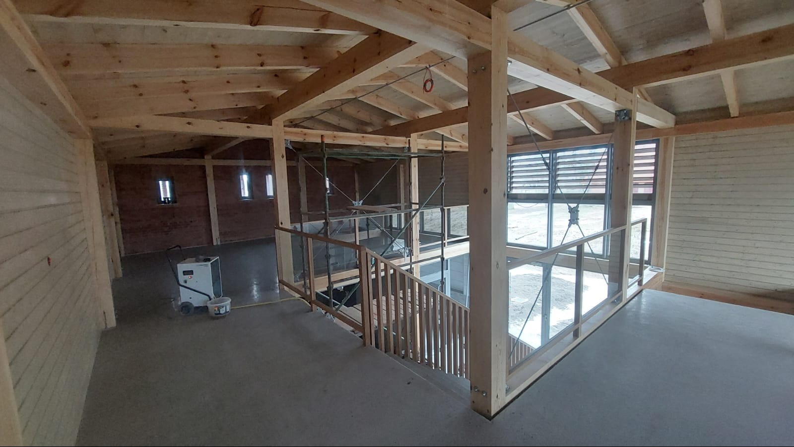 Wnętrze domu szkieletowego w trakcie budowy: drewniana konstrukcja, schody z balustradą, rusztowania, maszyna budowlana. Widoczne okna i drewniane wykończenie ścian.