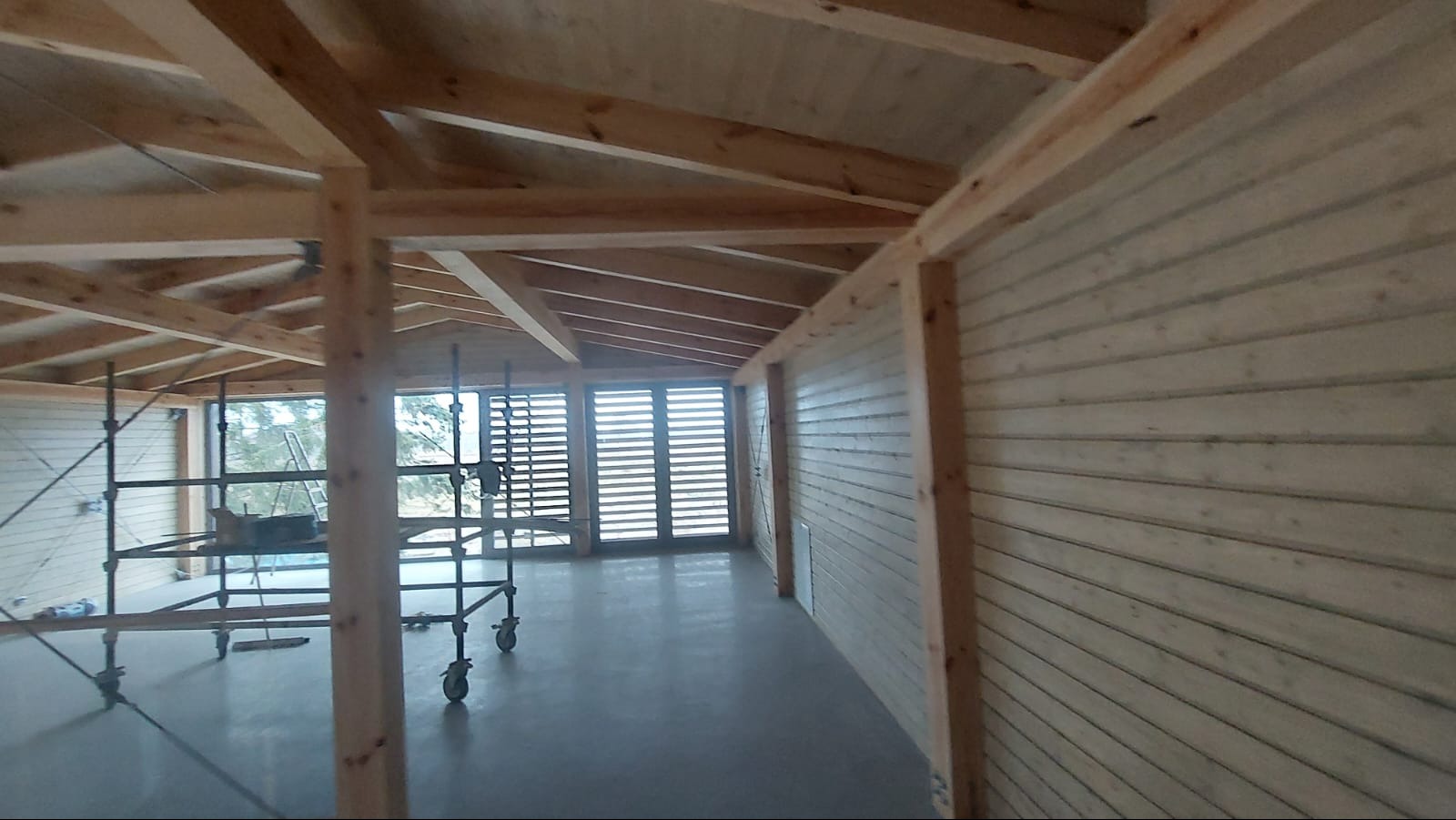 Wnętrze drewnianej konstrukcji w trakcie budowy, widoczne rusztowania, drewniane ściany z desek i elementy więźby dachowej. Jasne, naturalne oświetlenie.