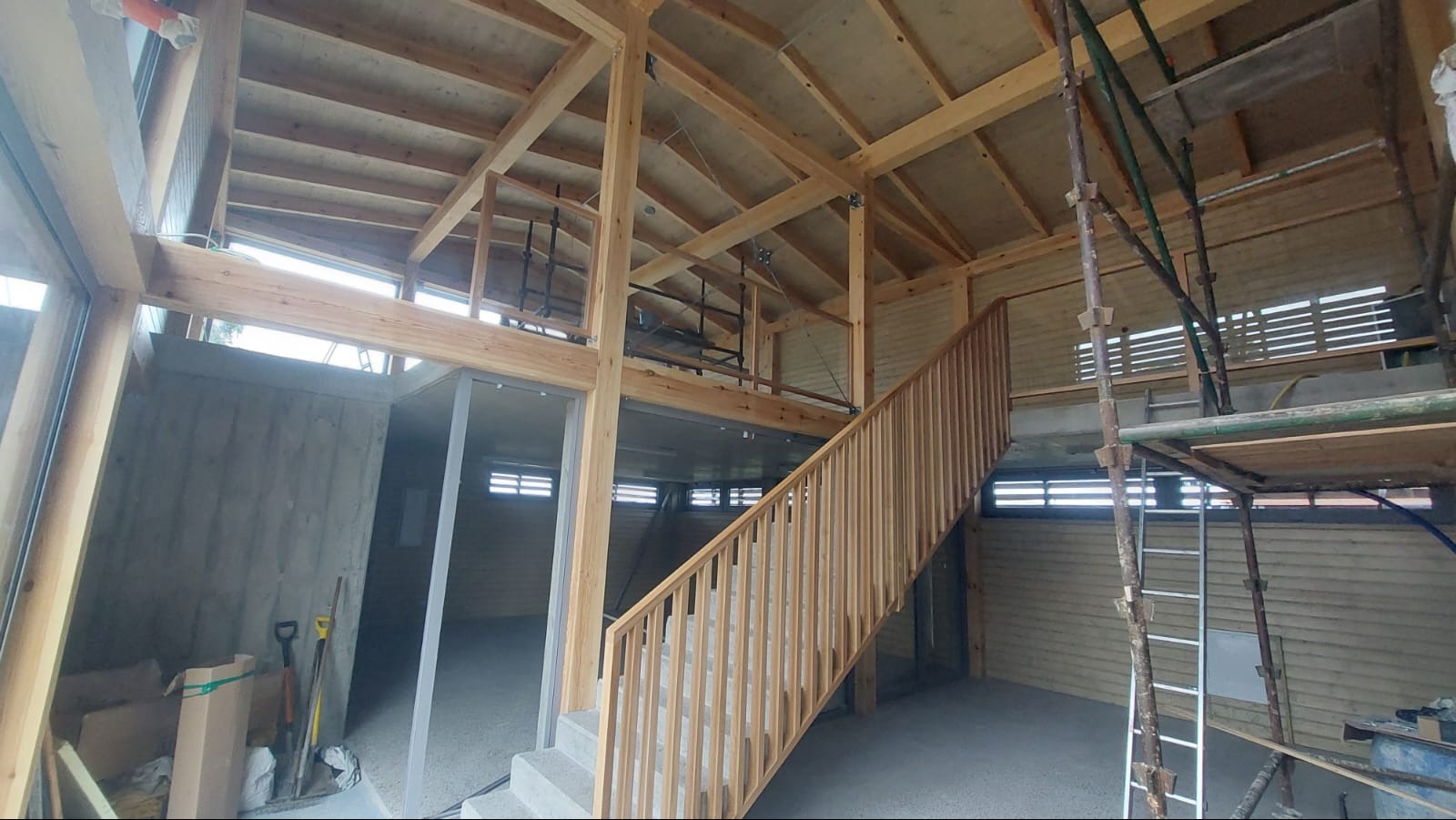 Wnętrze domu w trakcie budowy z widoczną drewnianą konstrukcją szkieletową, schodami i elementami betonowymi. Widoczne rusztowania i narzędzia budowlane.