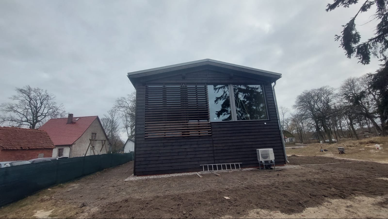 Czarny, drewniany dom szkieletowy z żaluzjami i dużym oknem, widoczny z zewnątrz na tle pochmurnego nieba i drzew. Na ziemi leży drabina.