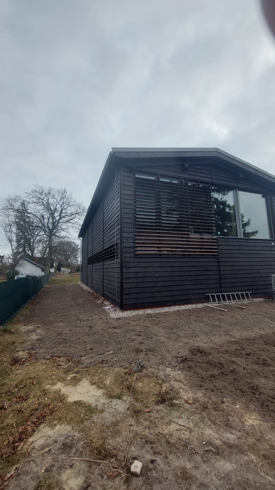 Czarny dom szkieletowy z drewnianymi żaluzjami i drabiną opartą o ścianę, widok z zewnątrz na tle pochmurnego nieba i drzew bez liści.