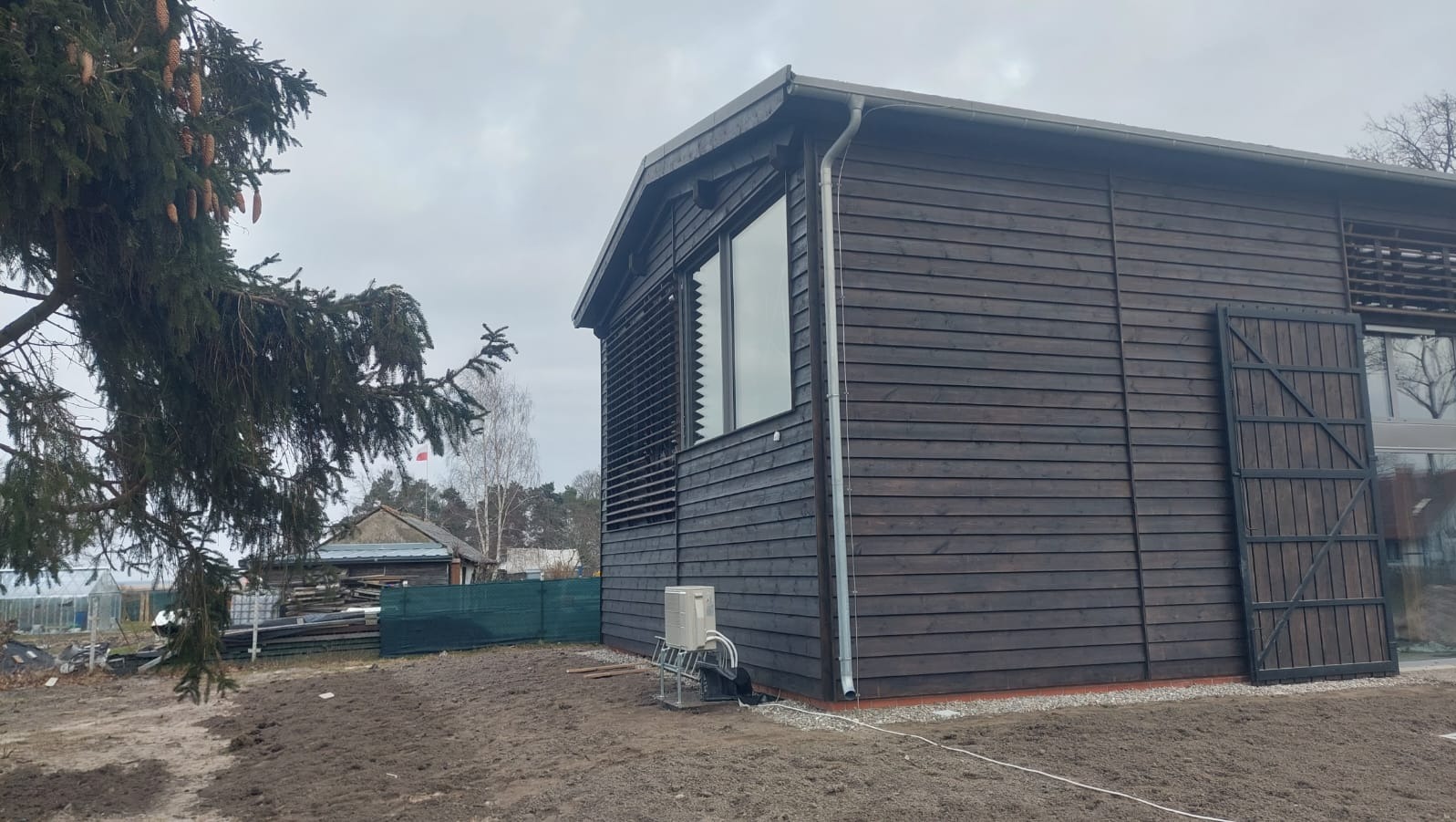 Ciemny, drewniany dom szkieletowy z widocznymi poziomymi deskami elewacyjnymi, dużym oknem z żaluzjami i metalowymi drzwiami. Na zewnątrz widoczny klimatyzator.