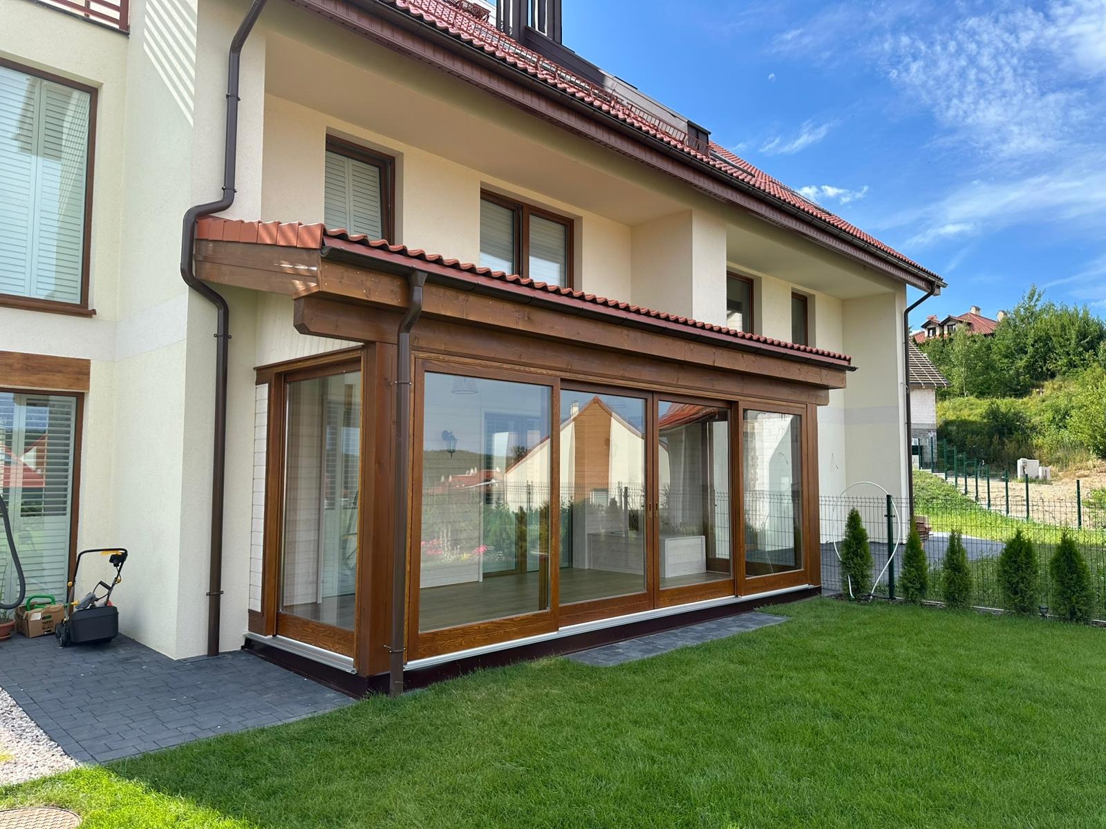 Nowoczesna, przeszklona weranda z drewnianą konstrukcją, przylegająca do jasnego domu z brązowym dachem. Widok z zewnątrz na zadbany trawnik i elementy architektury.