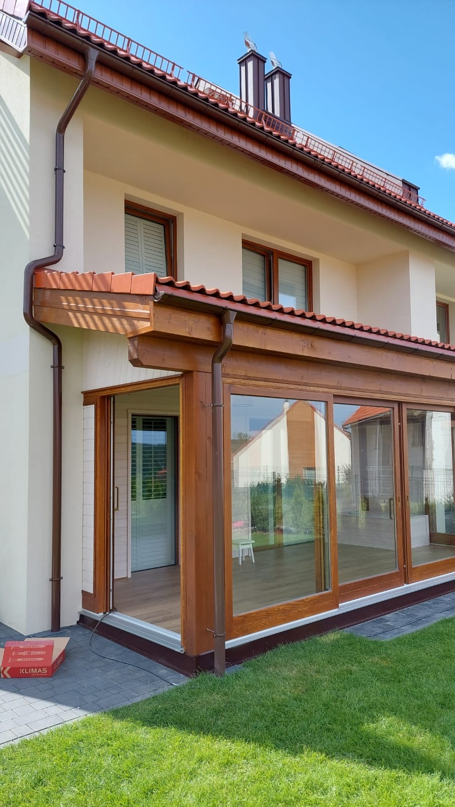 Zadaszony taras z drewnianą konstrukcją i przesuwnymi szklanymi drzwiami, brązowe rynny, widok z zewnątrz na zadbany trawnik i kostkę brukową.