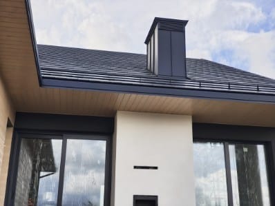 Nowoczesny dom z ciemną dachówką i metalowym kominem. Widoczne okna z roletami zewnętrznymi i drewniana podbitka dachowa. Ujęcie z dołu, podkreślające geometrię budynku.