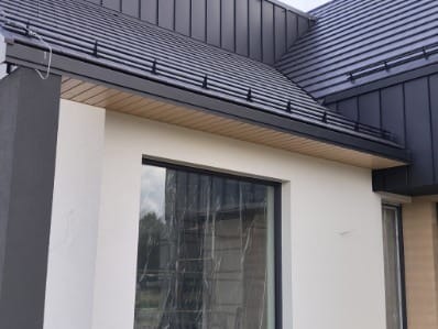 Nowoczesny dom z ciemną, metalową dachówką i jasną elewacją. Widoczny fragment okna z folią ochronną i drewniane podbitki dachowe.