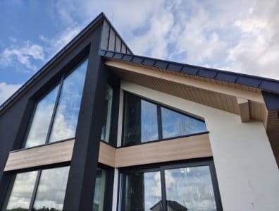 Nowoczesny dom szkieletowy z dużymi oknami i ciemną elewacją w Gdańsku. Widok z dołu, podkreślający geometrię budynku i odbicie chmur w szybach.