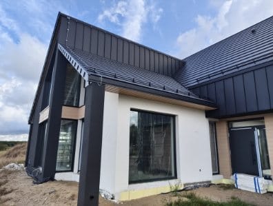Nowoczesny dom szkieletowy z ciemną blachą na dachu i dużymi oknami, widok z zewnątrz, częściowo wykończony, z widocznym ociepleniem fundamentów.