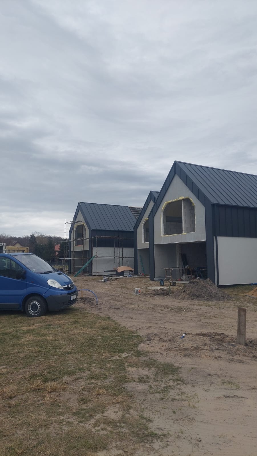 Dwa domy szkieletowe w trakcie budowy, z widoczną konstrukcją i czarnym dachem, na tle pochmurnego nieba. Na pierwszym planie zaparkowany niebieski samochód dostawczy.