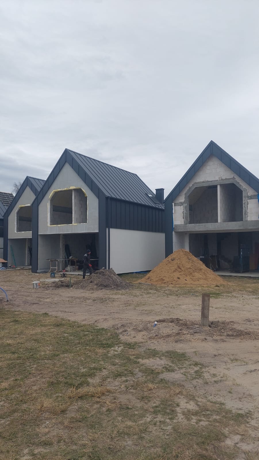 Surowe domy w zabudowie szeregowej z ciemnymi dachami z blachy na etapie budowy, widoczne sterty piasku i pracownik w oddali.
