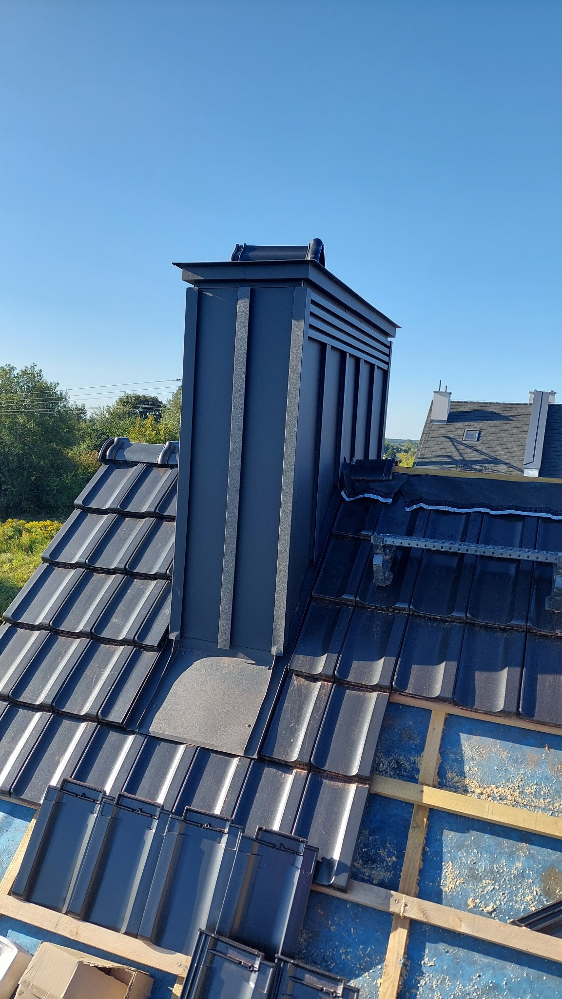Nowoczesny komin wentylacyjny z blachy na dachu krytym ciemną dachówką ceramiczną, widoczne elementy konstrukcji dachu i błękitne niebo w tle.