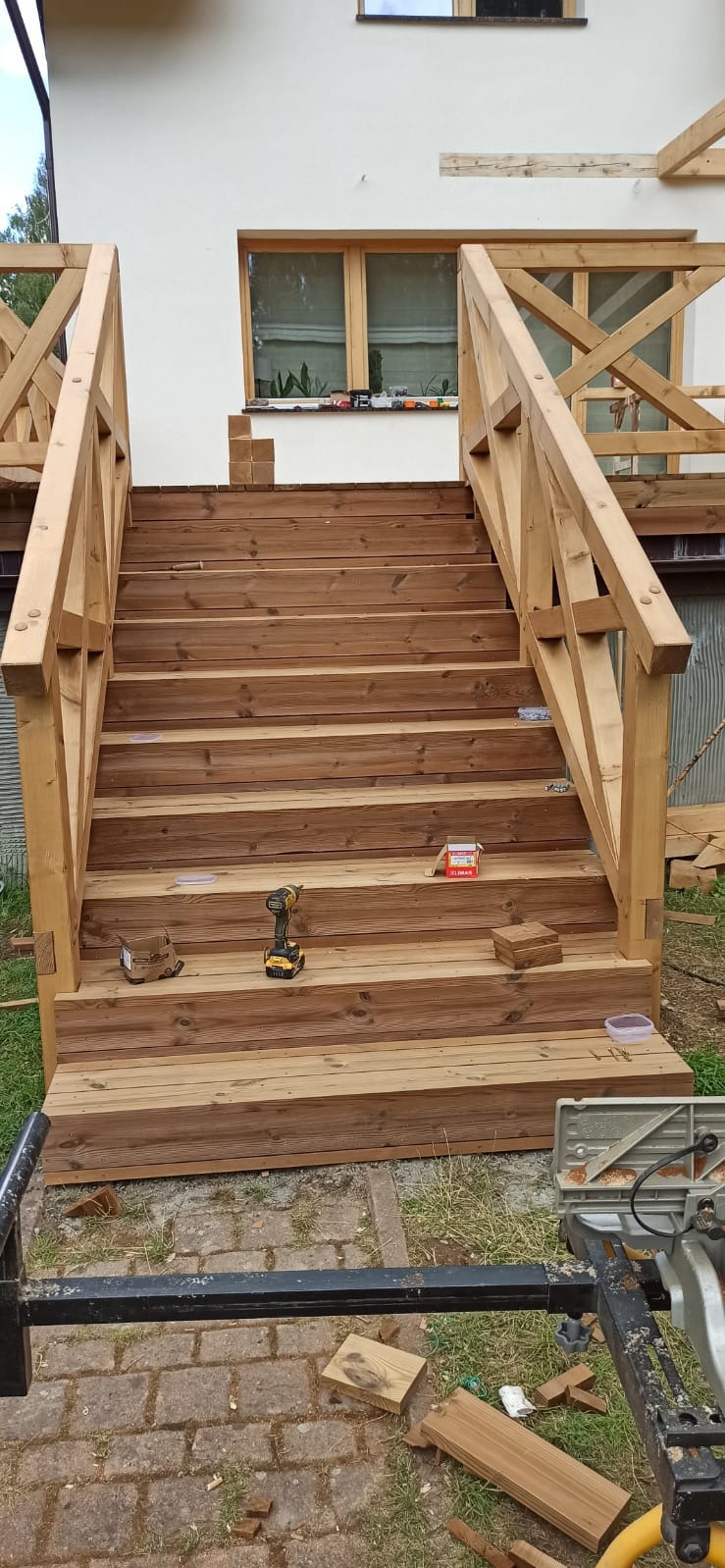 Drewniane schody z balustradą w trakcie budowy, widoczne narzędzia i materiały. Jasne drewno, dom w tle. Ujęcie z dołu, podkreślające konstrukcję schodów.