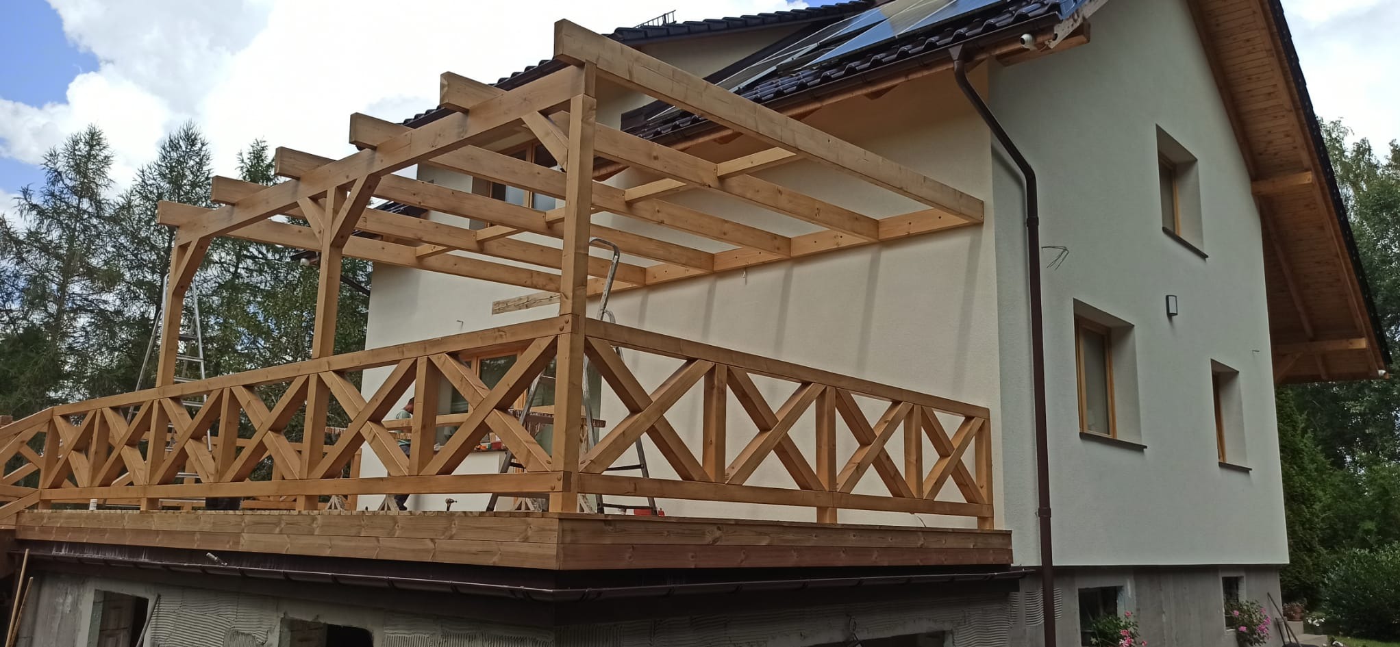 Drewniana konstrukcja tarasu z balustradą w trakcie budowy, widoczna drabina oparta o ścianę domu. Jasne drewno, częściowo zadaszona konstrukcja.