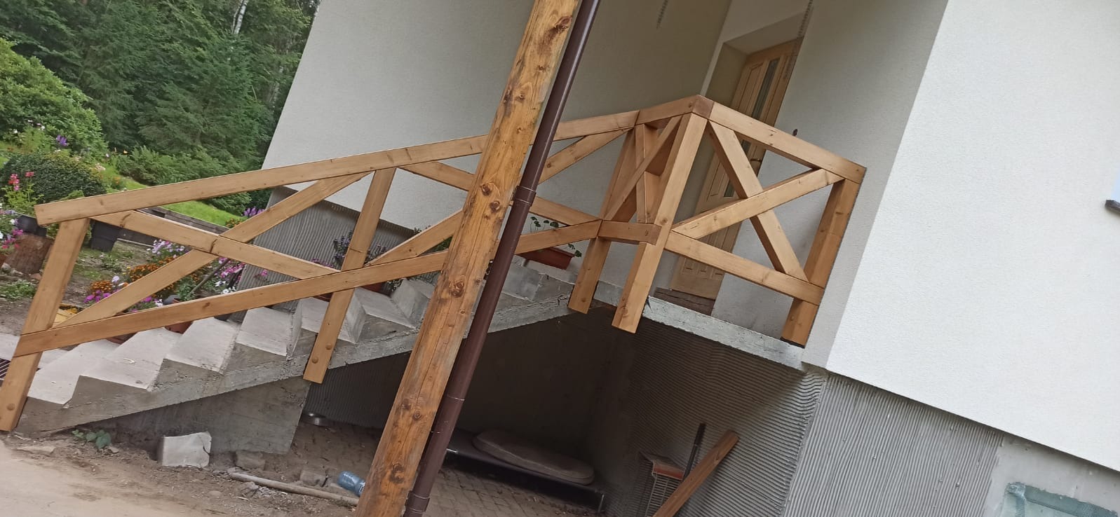 Drewniana balustrada schodów zewnętrznych z surowego drewna, z brązową rynną. W tle zieleń ogrodu i fragment elewacji budynku.