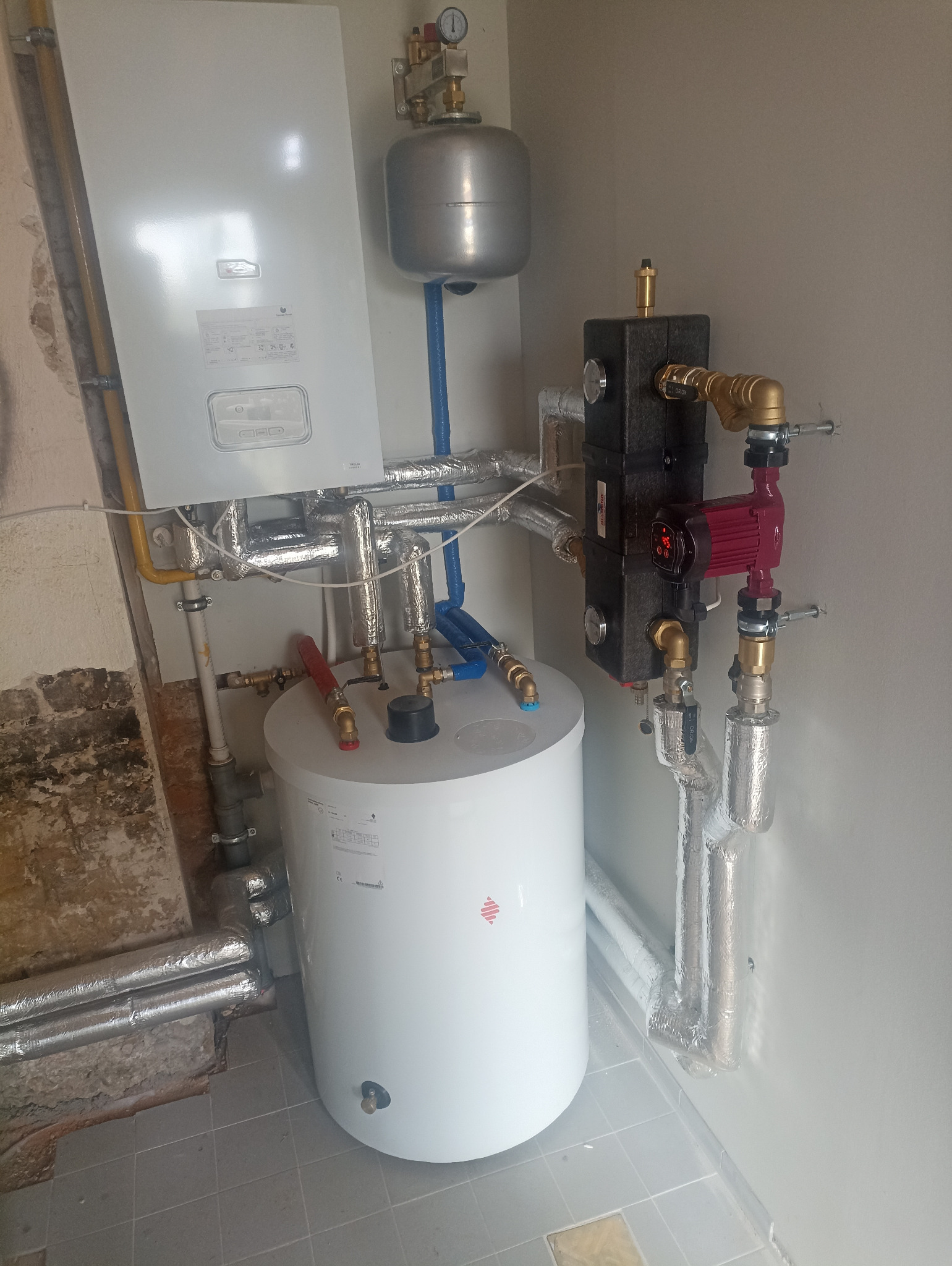 Kotłownia z zamontowanym kotłem gazowym Saunier Duval, widoczne rury w izolacji, zbiornik wyrównawczy i pompa obiegowa. Instalacja grzewcza w pomieszczeniu.