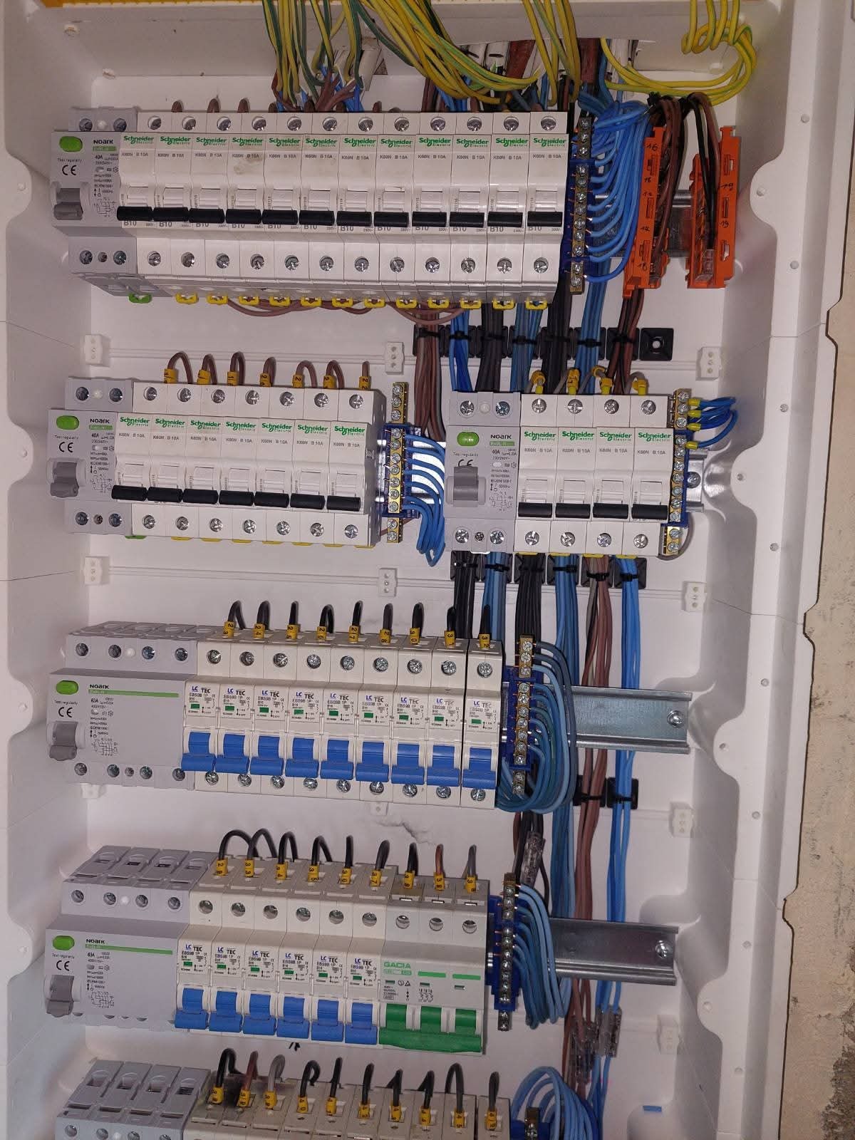 Rozdzielnica elektryczna z zamontowanymi bezpiecznikami, przewodami i listwami zaciskowymi. Widoczne różne kolory przewodów i elementy montażowe wewnątrz białej obudowy.