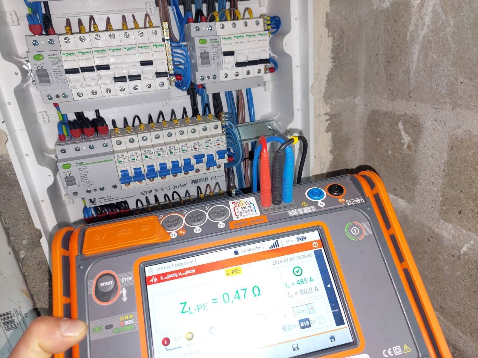 Rozdzielnica elektryczna z zamontowanymi bezpiecznikami, widoczny miernik parametrów instalacji z wyświetlanym wynikiem pomiaru impedancji pętli zwarcia.