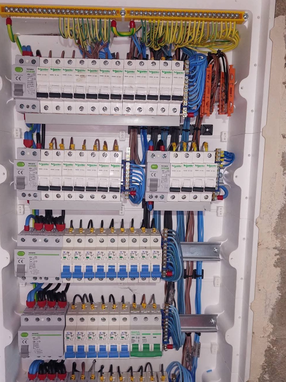 Rozdzielnica elektryczna z licznymi zabezpieczeniami, przewodami w różnych kolorach i listwami zaciskowymi, zamontowana w białej obudowie na tle surowej ściany.