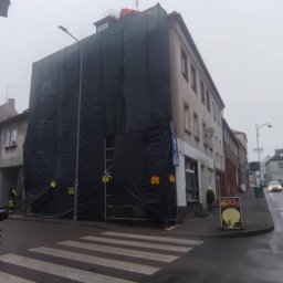 PRZEDSIĘBIORSTWO HANDLOWO-USŁUGOWE HUB-CRAFFT Hubert Wszelaki - Budynek w rusztowaniach, osłonięty czarną folią, prawdopodobnie w trakcie prac remontowych. Widoczne fragmenty dachu i elewacji. Ulica miejska, pochmurny dzień.